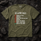 An olive green graphic t-shirt laid flat on a dark, textured rock background. The shirt features a white hand-written style text graphic reading 'MY GUITAR SKILLS:' followed by a checklist with red and green checkmarks next to various guitar-related phrases.