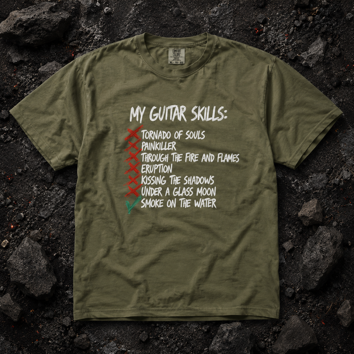 An olive green graphic t-shirt laid flat on a dark, textured rock background. The shirt features a white hand-written style text graphic reading 'MY GUITAR SKILLS:' followed by a checklist with red and green checkmarks next to various guitar-related phrases.