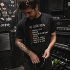 A man with tattoos wearing a black graphic t-shirt in a guitar amplifier room. The t-shirt features a white hand-written style list titled 'MY GUITAR SKILLS:' with 'Tornado of Souls', 'Pantherkiller', 'Through the Fire and Flames', 'Eruption', 'Kissing the Shadows', 'Under a Glass Moon', and 'Smoke on the Water' written below, with the first four crossed out in red and the last one checked in green.