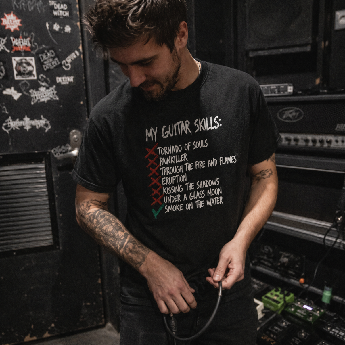 A man with tattoos wearing a black graphic t-shirt in a guitar amplifier room. The t-shirt features a white hand-written style list titled 'MY GUITAR SKILLS:' with 'Tornado of Souls', 'Pantherkiller', 'Through the Fire and Flames', 'Eruption', 'Kissing the Shadows', 'Under a Glass Moon', and 'Smoke on the Water' written below, with the first four crossed out in red and the last one checked in green.