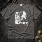 A dark gray graphic t-shirt laid flat on a textured dark background, featuring a white print of a portrait and the text 'LARS WAS RIGHT ABOUT NAPSTER (maybe)'. Studded leather and gold records are styled around the shirt.