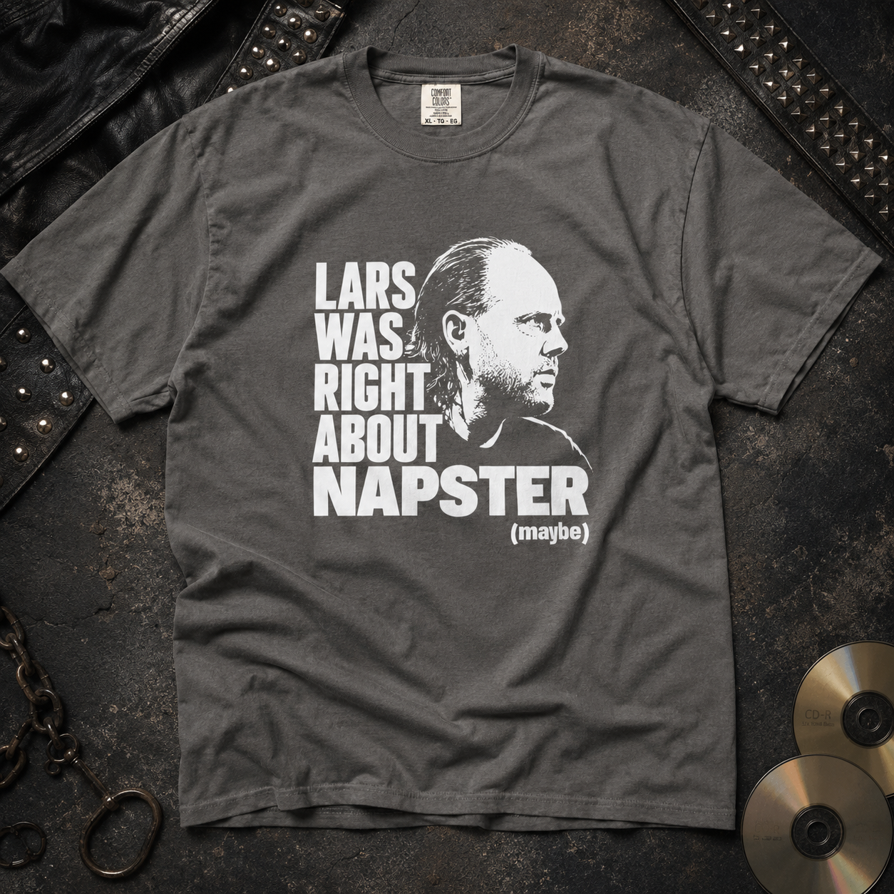 A dark gray graphic t-shirt laid flat on a textured dark background, featuring a white print of a portrait and the text 'LARS WAS RIGHT ABOUT NAPSTER (maybe)'. Studded leather and gold records are styled around the shirt.