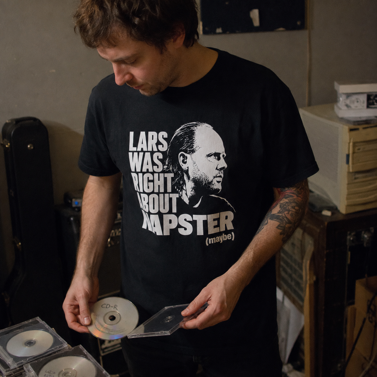 A man wearing a black graphic t-shirt with a white print of a portrait and the text 'LARS WAS RIGHT ABOUT RAPSTER (maybe)'. He is holding CD cases in a cluttered room.